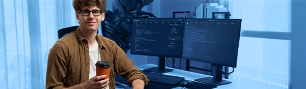 A programmer at a workstation with two monitors, holding a reusable coffee cup and looking forward.